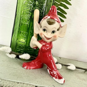 May include: A ceramic elf figurine wearing a red suit and pointed hat, holding a small beige baseball bat. The elf has large pointed ears, a smiling expression, and visible paint wear on the suit and hat. The figurine is standing upright.