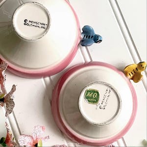 May include: Two ceramic bowls with pink exteriors and white interiors. One bowl features a blue insect-shaped handle, and the other has a yellow insect-shaped handle. The bases are stamped with MENSCHIK-GOLDMAN, INC. and one has a green M.G. JAPAN sticker.
