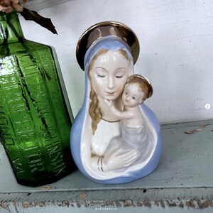 May include: A vintage ceramic figurine of Mary holding baby Jesus. The figurine features a blue robe, a gold halo, and delicate facial features. A green glass bottle is visible in the background.