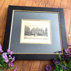 May include: A square black wooden picture frame with a thin gold inner border containing a black-and-white print of a city square with a large building, vintage cars, and pedestrians. The frame features a dark gray mat surrounding the central image.