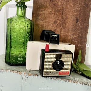 May include: A vintage Polaroid Swinger Model 20 Land Camera with a black and gold face, red shutter button, and a black viewfinder. A green glass bottle with text is to the left.