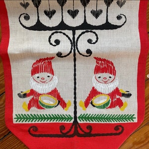 May include: A festive table runner with a red border and a beige center. The design features two gnomes in red outfits with white beards, holding drums. A black decorative element with hearts and a green leaf border complete the design.