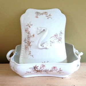 May include: Antique white porcelain wash basin set. The set includes a rectangular basin with handles and a matching pitcher with a swan-shaped handle. Floral and vine patterns in brown and pink decorate the set. A vintage bathroom decor item.
