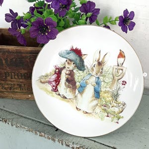 May include: A white ceramic plate with a gold rim featuring a colorful illustration of Peter Rabbit and friends. The design includes rabbits, a bird, and garden elements. The plate is surrounded by purple flowers.