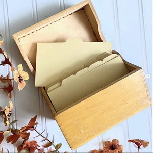 May include: A light-colored wooden recipe box with a hinged lid, featuring box joint construction on the corners. Inside the open box are several blank, cream-colored cardstock index dividers with rounded tabs, arranged in a vertical row.