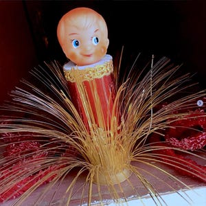 May include: A decorative figurine featuring a doll head with blue eyes and blonde hair atop a cylindrical red base. The base is wrapped in gold metallic trim and surrounded by a spray of thin, radiating gold tinsel-like fibers.