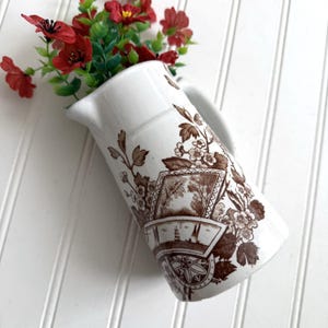 May include: A white ceramic pitcher featuring a brown transferware pattern of floral motifs, trees, and a nautical scene with sailboats. The pitcher is filled with a cluster of artificial red flowers with green stems and leaves.