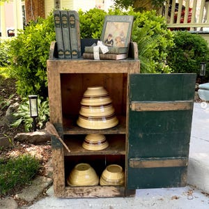 May include: A rustic wooden cabinet with a dark green door, displaying vintage items. The cabinet holds several yellow and brown striped mixing bowls, books, and a decorative book. The cabinet is outdoors, surrounded by greenery.