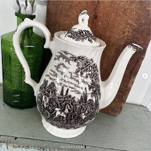May include: A white ceramic teapot with a black and white floral design. The teapot has a lid and a spout. The design features a horse-drawn carriage and trees.