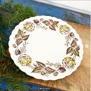 May include: A round, white ceramic plate with a scalloped edge, featuring a decorative floral wreath pattern. The design includes yellow and purple roses, small blue flowers, and detailed brown leaves arranged in a circular border.