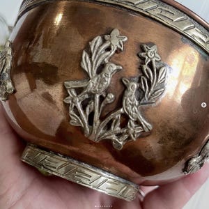 May include: A polished copper bowl featuring a silver-toned metal relief of two birds perched among flowers. The bowl has a wide silver-toned band around the rim and a matching silver-toned base, both decorated with a repeating geometric pattern.