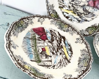 May include: A set of antique cream-colored plates with a scenic illustration of a winter landscape featuring a red-roofed building. The plates have a scalloped edge with a brown and green floral border.