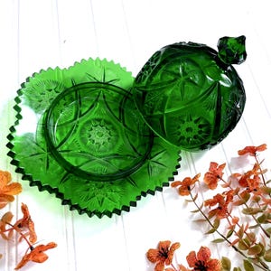 May include: A green glass serving dish set with a starburst pattern. The set includes a round bowl with a lid and a square plate with a scalloped edge. The glass has a rich, emerald green color. Decorative orange flowers are in the foreground.