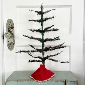May include: A vintage-style Christmas tree with a green wire frame and dark green, bristly branches. The tree sits on a red fabric base with a white lace trim. The tree is displayed on a light blue, weathered wooden surface.