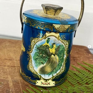 May include: A vintage blue metal container with a hinged lid and a handle. The container features a decorative gold floral design and a framed image of two yellow birds with long tail feathers. The container is approximately 6 inches tall.