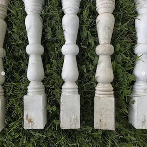 May include: A collection of white, weathered wooden balusters, each with a turned design featuring a square base, a central bulbous section, and a decorative top. The balusters are arranged on a bed of green grass.