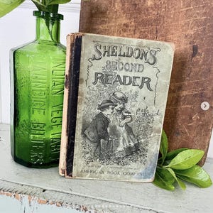 May include: An antique book titled "Sheldon's Second Reader" with a faded cover illustration of two children. A green glass bottle with text is to the left. The book is from the American Book Company.