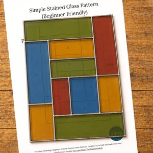 May include: A rectangular stained glass panel featuring a geometric arrangement of green, blue, yellow, and red glass segments held together by silver metal solder lines. The panel is placed over a paper document titled Simple Stained Glass Pattern.