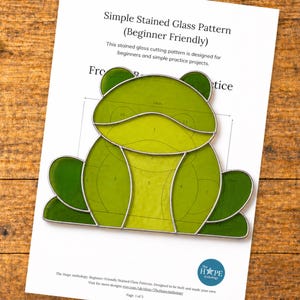 May include: A beginner-friendly stained glass pattern featuring a frog design. The pattern is printed on a white sheet of paper and includes the text "Simple Stained Glass Pattern (Beginner Friendly)". The frog is composed of various shades of green.