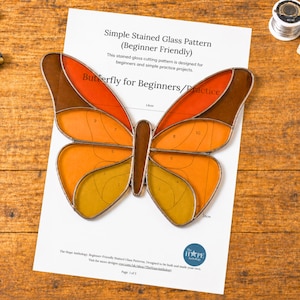 Puede incluir: Un patrón de vidriera de mariposa en tonos naranja, marrón y amarillo, sobre papel blanco con texto. El papel dice "Butterfly for Beginners/Practice" y "Simple Stained Glass Pattern (Beginner Friendly)." Se ven herramientas.