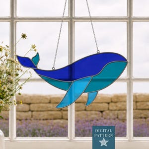 Peut inclure: Un attrape-soleil en vitrail en forme de baleine composé de pièces de verre dans des tons bleu et turquoise assemblées par des lignes de soudure en métal. La baleine est suspendue par deux fines chaînes argentées et porte une étiquette DIGITAL PATTERN.
