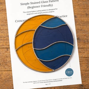 Op de afbeelding: Een beginnersvriendelijk glas-in-loodpatroon met een halve maan ontwerp. Het patroon gebruikt gele en blauwe glazen secties, omlijnd met zilver metaal. De tekst op het papier luidt: "Simple Stained Glass Pattern" en "Crescent Moon for Beginners Practice."