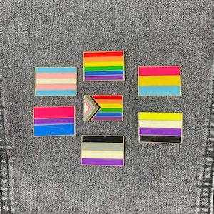 May include: A collection of enamel pins featuring various LGBTQ+ pride flags. The pins have a gold border and are set against a gray denim background. Flags include the rainbow, transgender, bisexual, pansexual, non-binary, and asexual flags.
