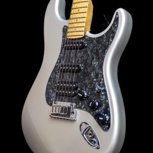 Pu&ograve; includere: Chitarra elettrica argentata con battipenna nero e manopole di controllo nere. La chitarra ha un manico in acero e un ponte cromato. Il corpo &egrave; di colore argento pieno e lo sfondo &egrave; nero.