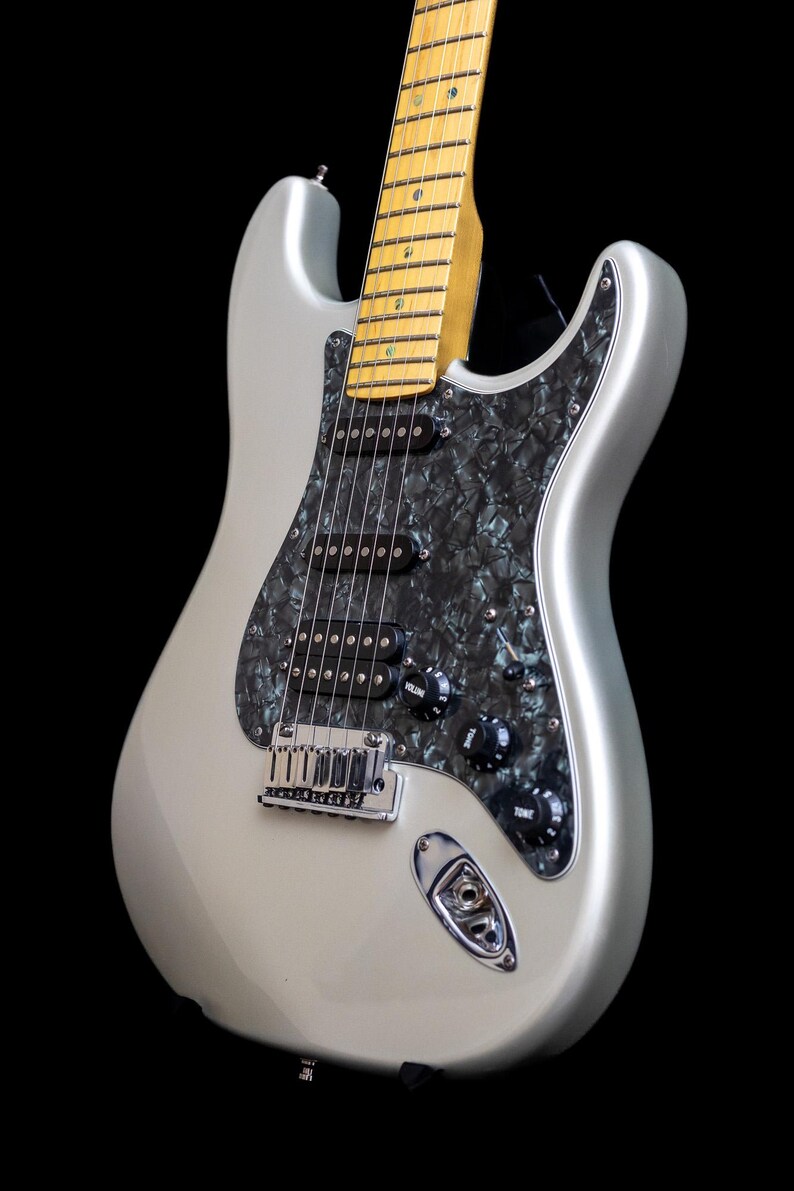 Pu&ograve; includere: Chitarra elettrica argentata con battipenna nero e manopole di controllo nere. La chitarra ha un manico in acero e un ponte cromato. Il corpo &egrave; di colore argento pieno e lo sfondo &egrave; nero.