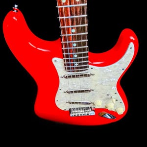 Pu&ograve; includere: Una chitarra elettrica rosso vivo con battipenna bianco e hardware cromato. La chitarra ha manico e tastiera in acero e manopole color crema. Lo strumento &egrave; ambientato su sfondo nero.