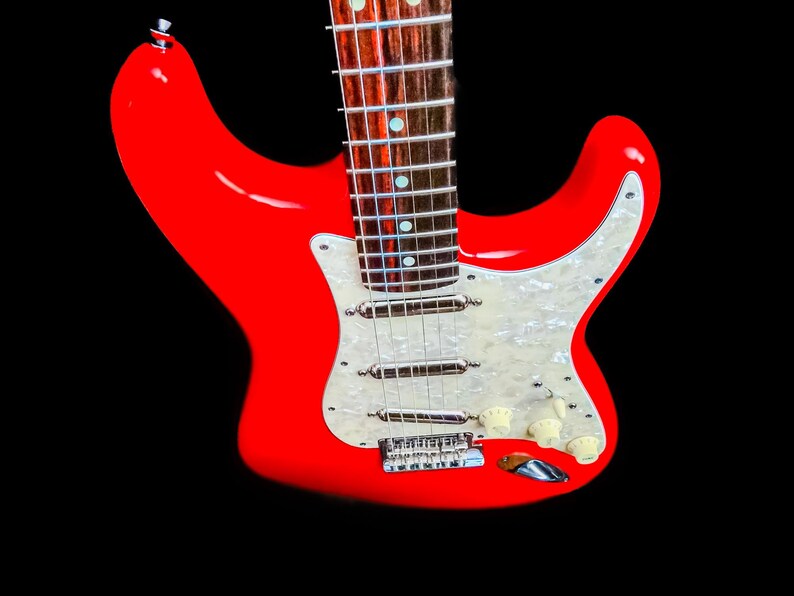 Pu&ograve; includere: Una chitarra elettrica rosso vivo con battipenna bianco e hardware cromato. La chitarra ha manico e tastiera in acero e manopole color crema. Lo strumento &egrave; ambientato su sfondo nero.