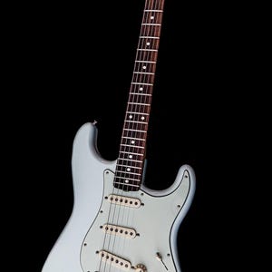 Pu&ograve; includere: Una chitarra elettrica azzurra con battipenna bianco e tre pickup. La chitarra ha manico e paletta in legno con meccaniche. Il corpo ha una forma classica, su sfondo nero.