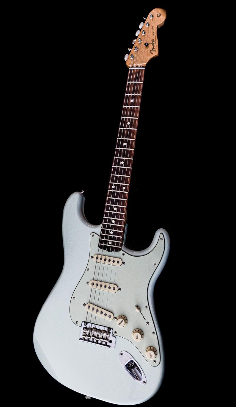 Pu&ograve; includere: Una chitarra elettrica azzurra con battipenna bianco e tre pickup. La chitarra ha manico e paletta in legno con meccaniche. Il corpo ha una forma classica, su sfondo nero.