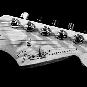 Pu&ograve; includere: Immagine in bianco e nero della paletta di una chitarra Fender Stratocaster. La paletta presenta meccaniche, corde e il logo Fender con la scritta "Corona, California". Sono visibili anche le parole "Stratocaster" e "Original Contour Body".