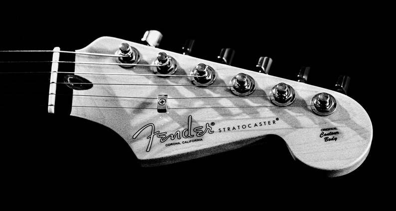 Pu&ograve; includere: Immagine in bianco e nero della paletta di una chitarra Fender Stratocaster. La paletta presenta meccaniche, corde e il logo Fender con la scritta "Corona, California". Sono visibili anche le parole "Stratocaster" e "Original Contour Body".