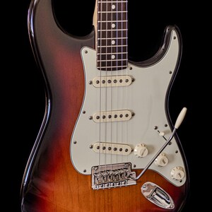 Pu&ograve; includere: Chitarra elettrica con finitura sunburst, battipenna bianco, tre pickup single-coil e hardware cromato. La chitarra presenta manico in acero e tastiera in palissandro, con manopole bianche e leva del tremolo.