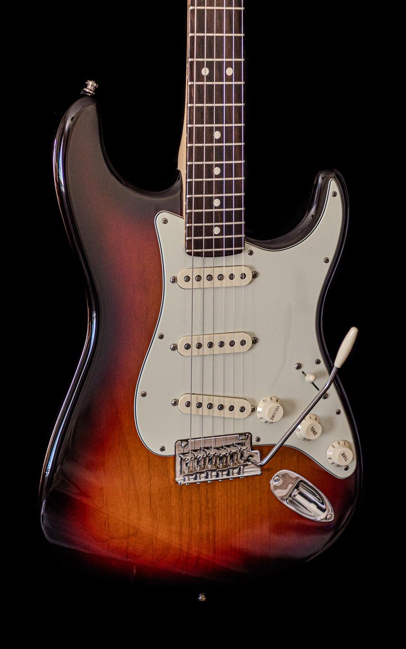Pu&ograve; includere: Chitarra elettrica con finitura sunburst, battipenna bianco, tre pickup single-coil e hardware cromato. La chitarra presenta manico in acero e tastiera in palissandro, con manopole bianche e leva del tremolo.