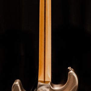 Pu&ograve; includere: Vista posteriore di una chitarra elettrica argento metallizzato su sfondo nero. La chitarra presenta un manico in acero con una striscia scura, meccaniche cromate e un battipenna nero. Il logo Fender &egrave; visibile.
