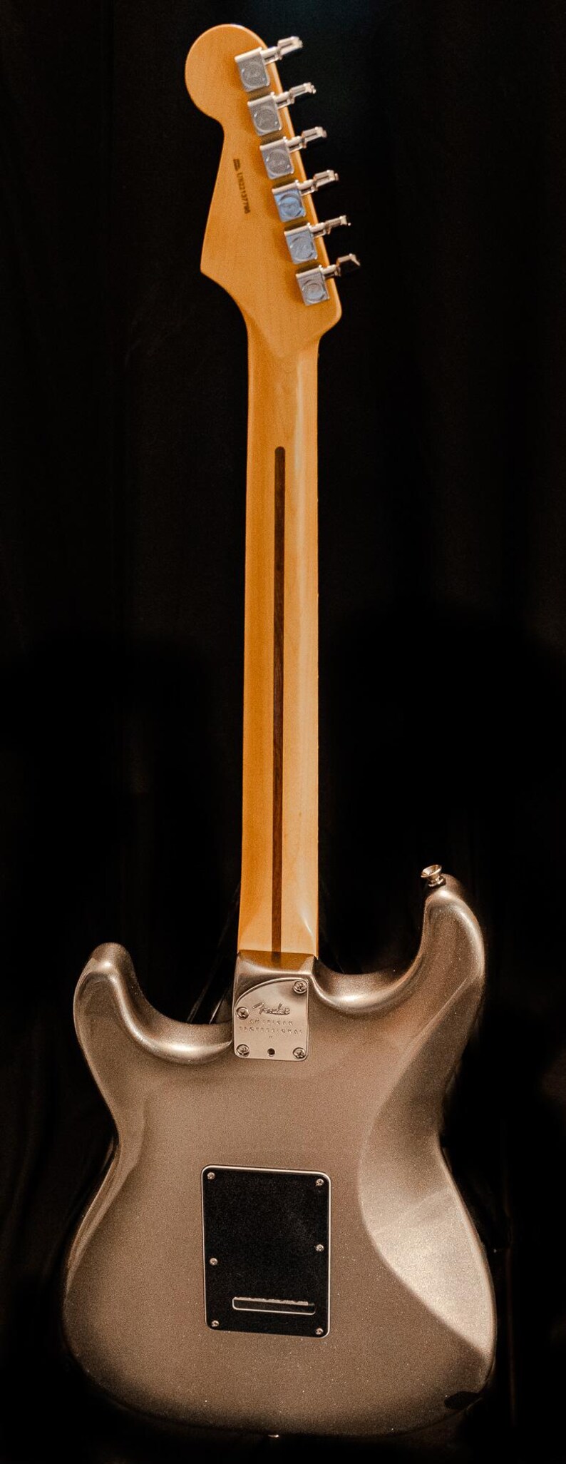Pu&ograve; includere: Vista posteriore di una chitarra elettrica argento metallizzato su sfondo nero. La chitarra presenta un manico in acero con una striscia scura, meccaniche cromate e un battipenna nero. Il logo Fender &egrave; visibile.