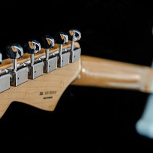 Pu&ograve; includere: Primo piano della paletta e del manico di una chitarra elettrica azzurra su sfondo nero. La paletta &egrave; in legno chiaro e presenta sei meccaniche cromate. Il manico &egrave; in legno chiaro e il corpo &egrave; azzurro.