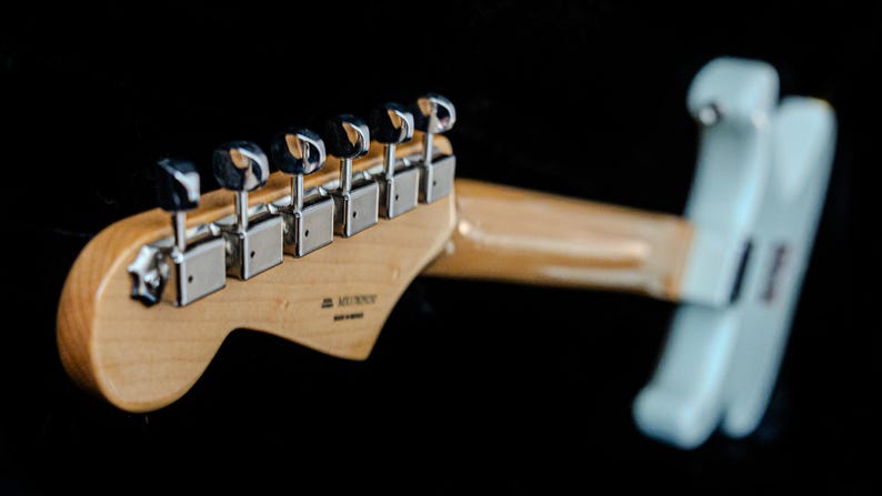 Pu&ograve; includere: Primo piano della paletta e del manico di una chitarra elettrica azzurra su sfondo nero. La paletta &egrave; in legno chiaro e presenta sei meccaniche cromate. Il manico &egrave; in legno chiaro e il corpo &egrave; azzurro.