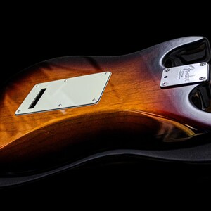 Pu&ograve; includere: Primo piano del retro di una chitarra elettrica con finitura sunburst. Il corpo ha una sfumatura di marrone, arancione e nero. Sono visibili un battipenna bianco e una placca argentata per il manico. La chitarra &egrave; su sfondo nero.