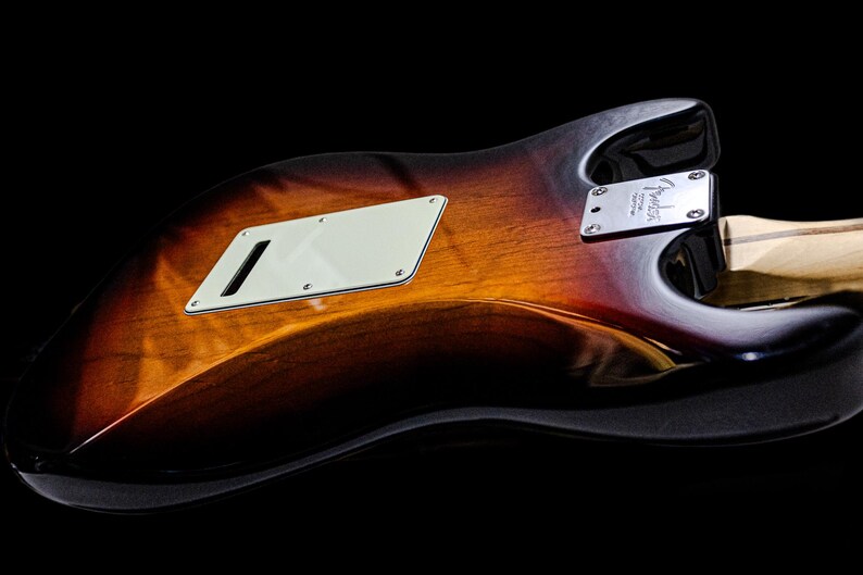 Pu&ograve; includere: Primo piano del retro di una chitarra elettrica con finitura sunburst. Il corpo ha una sfumatura di marrone, arancione e nero. Sono visibili un battipenna bianco e una placca argentata per il manico. La chitarra &egrave; su sfondo nero.