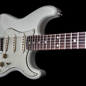 Pu&ograve; includere: Una chitarra elettrica grigio chiaro con tastiera marrone scuro e segnatasti bianchi. La chitarra ha tre pickup, manopole del tono e del volume e un ponte tremolo. Lo sfondo &egrave; nero.