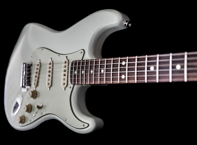 Pu&ograve; includere: Una chitarra elettrica grigio chiaro con tastiera marrone scuro e segnatasti bianchi. La chitarra ha tre pickup, manopole del tono e del volume e un ponte tremolo. Lo sfondo &egrave; nero.
