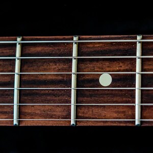 Pu&ograve; includere: Primo piano della tastiera di una chitarra. Il legno marrone scuro &egrave; contrassegnato da indicatori di tasti bianchi e tasti argentati. La venatura del legno &egrave; visibile e l'immagine &egrave; su sfondo nero.