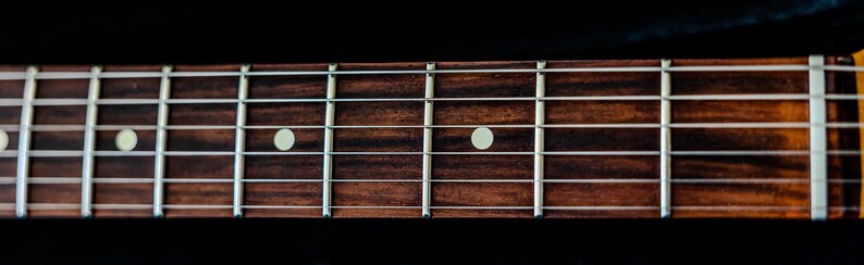 Pu&ograve; includere: Primo piano della tastiera di una chitarra. Il legno marrone scuro &egrave; contrassegnato da indicatori di tasti bianchi e tasti argentati. La venatura del legno &egrave; visibile e l'immagine &egrave; su sfondo nero.