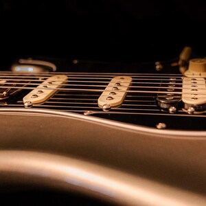 Pu&ograve; includere: Primo piano di una chitarra elettrica argentata con corde, pickup e manopole di controllo. La chitarra ha un battipenna nero e un manico in legno. L'immagine &egrave; scattata con una profondit&agrave; di campo ridotta, concentrandosi sulle corde e sui pickup.