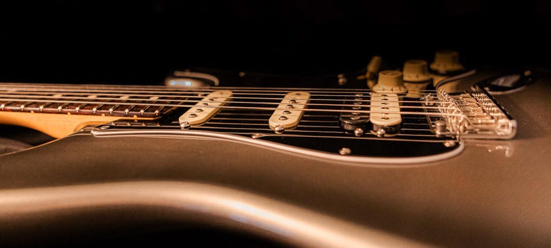 Pu&ograve; includere: Primo piano di una chitarra elettrica argentata con corde, pickup e manopole di controllo. La chitarra ha un battipenna nero e un manico in legno. L'immagine &egrave; scattata con una profondit&agrave; di campo ridotta, concentrandosi sulle corde e sui pickup.