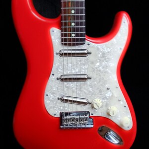 Pu&ograve; includere: Una vivace chitarra elettrica rossa con battipenna perlato. La chitarra presenta hardware cromato, inclusi pickup, ponte e meccaniche. Sono visibili manopole di controllo bianche. Il corpo della chitarra &egrave; rosso pieno e lucido.